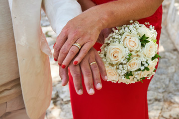 A couple holding a wedding bouquet in front of them, man in white and woman in red dress rings on fingers