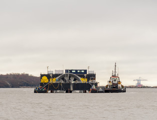 A giant tidal turbine being delivered for installation on the ocean floor. Turbine is held by a delivery ship that is being moved by a tugboat. ID has been removed from the boats. Sky is overcast. © madscinbca