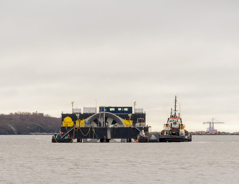 A giant tidal turbine being delivered for installation on the ocean floor. Turbine is held by a delivery ship that is being moved by a tugboat. ID has been removed from the boats. Sky is overcast.