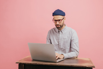 Concentrated hipster student reads information and checks data, focused in monitor of laptop...