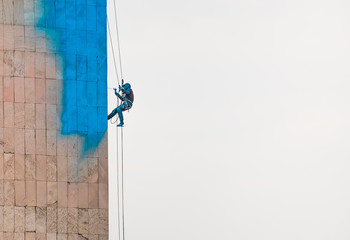 An industrial climber painting a building in vivid blue color hanging on a metal wire rope against highlighted sky background.