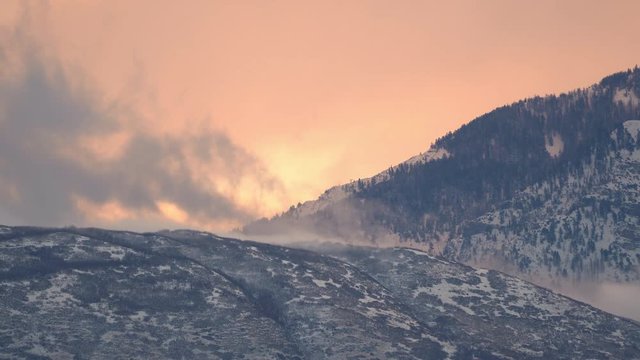 Low foggy clouds moving over snow covered mountains during colorful sunrise in Utah.