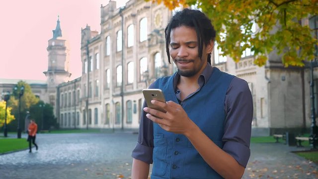 Portrait of African-American student with dreadlocks watching into smartphone changing emotions on university background.
