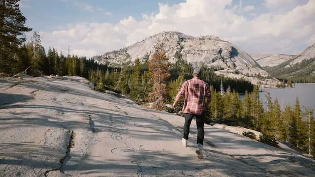 Camera follows young happy tourist man running down big rocks while hiking alone at Yosemite mountain lake slow motion.