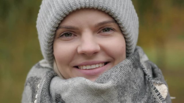 Portrait of young adorable woman in grey hat, scarf, blush jacket, standing on the wind, enjoying cold autumn day, smiling and looking into camera with fall colors background. Extreme close up shot.