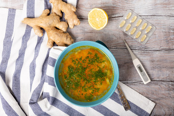 Health concept. Bowl of fresh homemade soup broth to cure flu, illness, sick. Lemon, thermometer, tablet in blister and ginger on white background, top view. Flat lay.