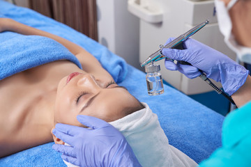 Crop master in gloves spraying cosmetic with airbrush on face of young Asian woman in spa salon