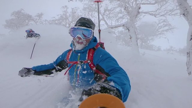 SLOW MOTION, SELFIE: Two cheerful male tourists having fun skiing in the untouched mountain terrain in Japan. Freeride skiers enjoying their winter vacation in Niseko by shredding fresh powder snow.