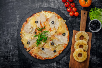 Pizza with chicken, pineapple, cheese and olives on stone and a black wooden scratched background