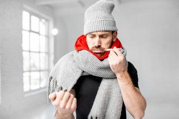 Portrait of a man wrapped in a warm scarves and hat feeling bad measuring body temperature indoors....