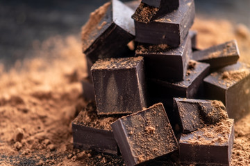 Pieces of dark bitter chocolate with cocoa powder on dark wooden background. Concept of confectionery ingredients