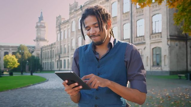 Portrait of attractive African-American student with dreadlocks working with tablet on university background.