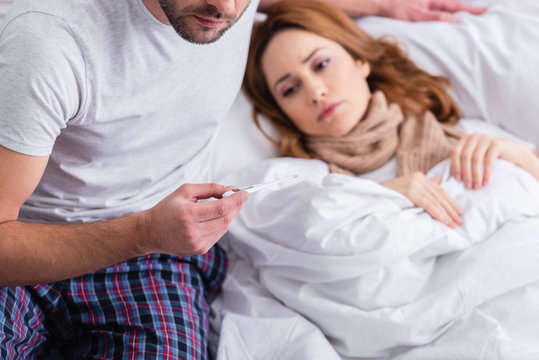cropped image of husband checking temperature of sick wife and holding thermometer in bedroom