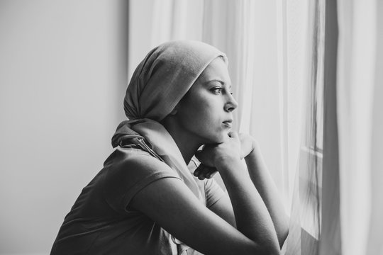 Black and white photo of thoughtful young girl suffering from ovarian cancer wearing headscarf and looking through the window in medical center