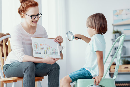 Smiling therapist showing picture of house to boy with magnifier during psychotherapy