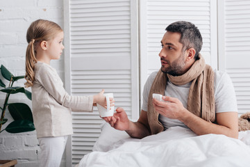 Fototapeta premium side view of adorable daughter giving cup of tea to sick dad in bedroom