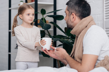 Obraz na płótnie Canvas adorable daughter giving cup of tea to sick dad in bedroom