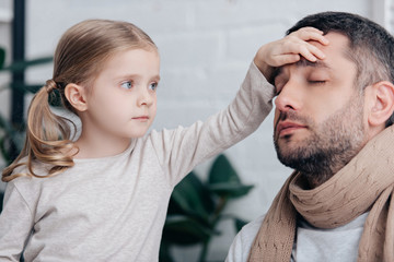 portrait of adorable daughter touching sick father forehead and checking temperature in bedroom