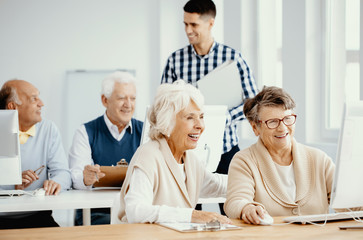 Group of active elderly people learning from young teacher how to use computer at third age university classes - obrazy, fototapety, plakaty