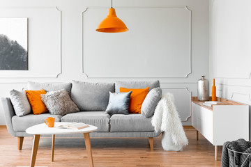 Orange lamp above table and grey settee with cushions in bright flat interior with poster. Real...