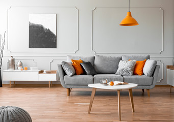 Orange lamp above wooden table in front of grey sofa in living room interior with poster. Real photo
