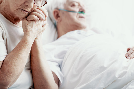 Sad senior woman holding hand of her cancer sick husband lying in hospice bed, photo with copy space