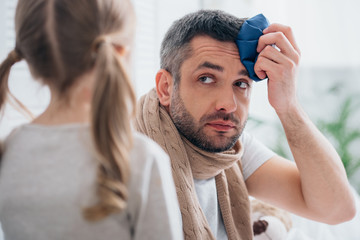 daughter looking at sick father touching head with ice pack in bedroom