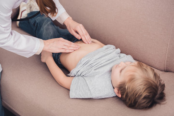 cropped image of pediatrist in white coat palpating sick boy stomach on sofa in living room
