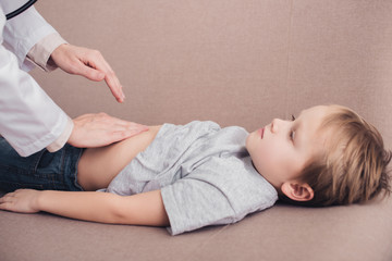 Obraz na płótnie Canvas cropped image of pediatrist palpating sick boy stomach on sofa in living room