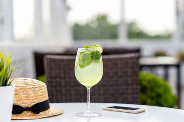 Close-up cafe table in summer in fresh air. On table lies a straw hat, a mobile phone, a wine glass with soft drinks of mint and lemon. The concept of ordering in Restaurant.