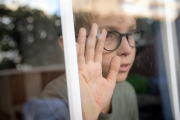 blond child with glasses, he looks calm and serene through the window with his hand resting on the glass. Lights of the sunset are reflected on the glass. Wait for friends to come play with him