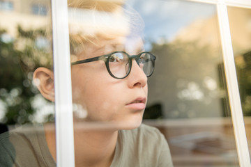 blond child with glasses, he looks calm and serene through the glass of the window, the lights of the sunset are reflected on the glass. Wait for friends to come play with him