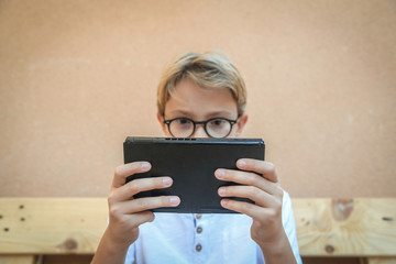 Boy on wooden bench in garden, playing absorbed with his console. Teenager engaged in favorite pastime. face with glasses behind a video game console. Employee videogame. Modern life in the open air