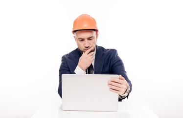 A tired constructor businessman in an orange helmet looks at the laptop screen and studies the construction project. Sits at the table and drinks coffee, nervous