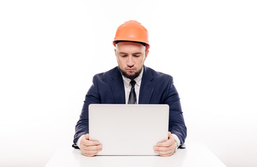 A tired constructor businessman in an orange helmet looks at the laptop screen and studies the construction project. Sits at the table and drinks coffee, nervous
