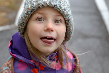 Girl posing with a cute face, showing tongue. Girl in a hat with a pompon, having fun on the street