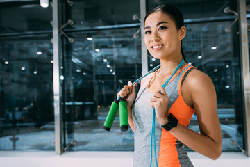 smiling asian sportswoman holding skipping rope on shoulders at gym