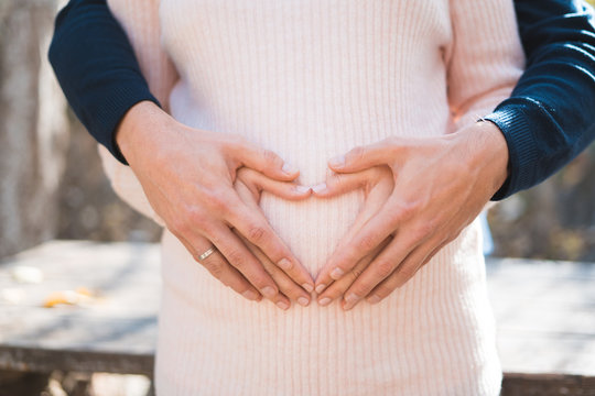 Cropped image of beautiful pregnant woman and her handsome husband hugging and making heart on the tummy.