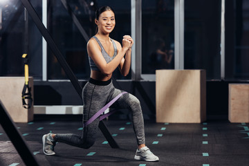 Obraz na płótnie Canvas smiling asian girl doing lunges with rubber resistance bands at gym