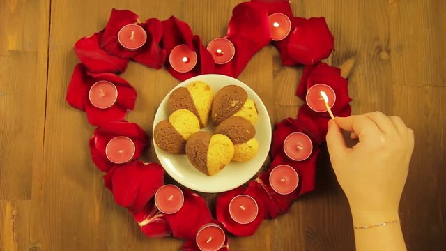 A girl lays out a heart on a wooden table from rose petals, places and lights candles. Time laps.