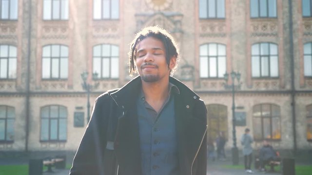 Portrait of contented African-American student with dreadlocks walking in sunshine from university and greeting someone.