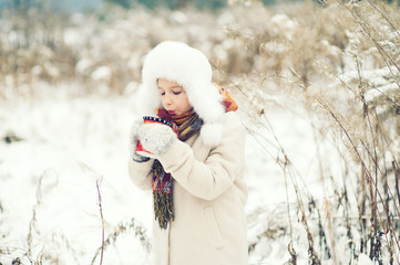 Beautiful russian toddler girl wearing furry hat drink hot beverage in snowy cold winter forest