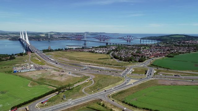 Aerial footage of traffic on the M9 motorway approaching and driving over the Queensferry Crossing bridge on the Firth of Forth with the old Forth Road Bridge and Railway Bridge beyond.