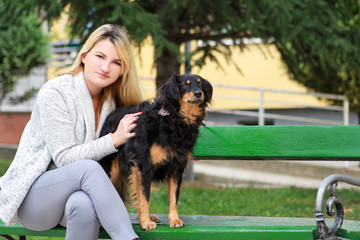 Beautiful woman with his small mixed breed dog sitting and posing in front of camera on wooden bench at city park. Portrait of owner and cute half breed dog enjoys, resting, petting together outdoors.