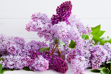 Purple flowers of lilac with leaves in a glass bottle on a white wooden background