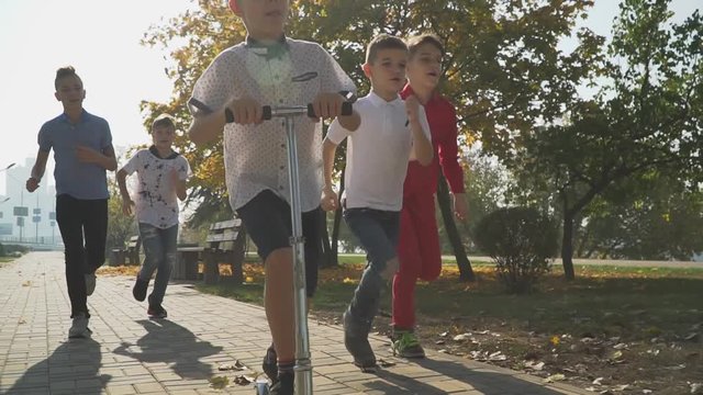 Kid rides a scooter in the autumn park. Friends are catching up with a boy riding a scooter outdoors. Kids playing in the park.