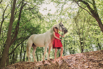 Young woman in red dress with her white horse in the forest