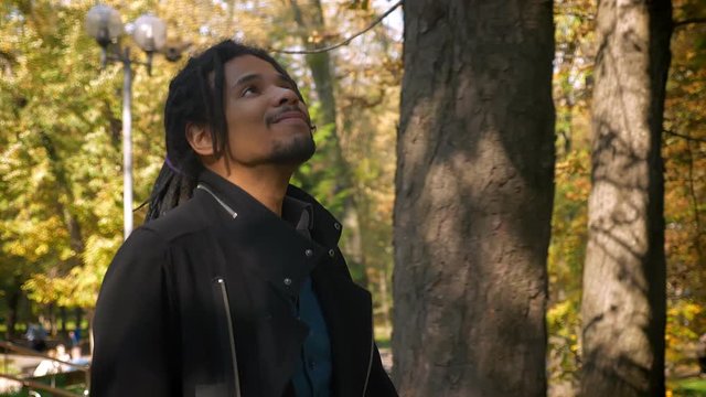 Portrait of African-American guy with dreadlocks walking around autumnal park.