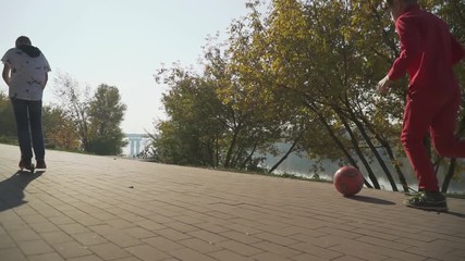 The boy is riding a scooter. Kid is running in the park kicking red ball