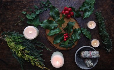 Yule winter solstice (Christmas) themed flat lay with branch of holly plant on a dark wooden table....
